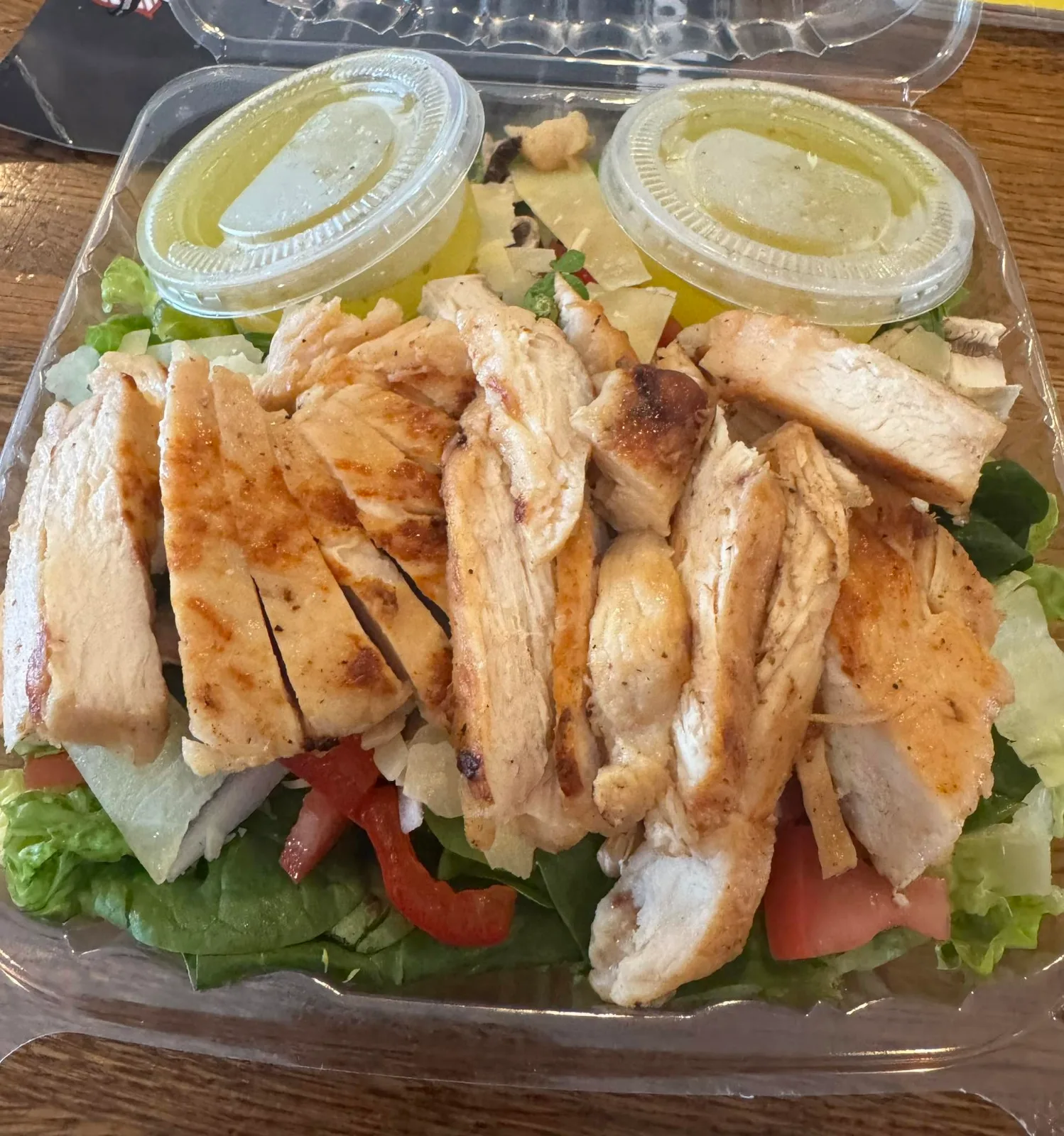 A grilled chicken salad in a takeout container with two tubs of dressing