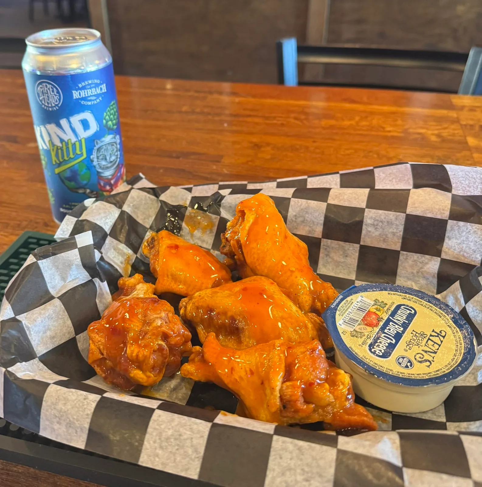 A basket of wings in hot sauce on checkered paper with a tub of blue cheese, beside a can of Three Heads and Rohrbach beer
